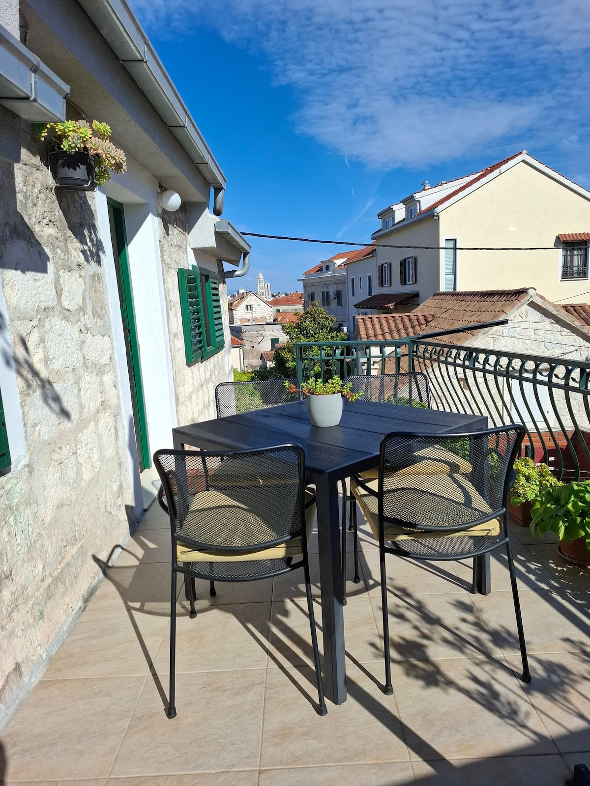 A patio area is shown, featuring a dark table with two mesh chairs. Small potted plants adorn the railing, while the picturesque Old Town is visible in the background. The sky is clear, enhancing the inviting ambiance of the outdoor space.