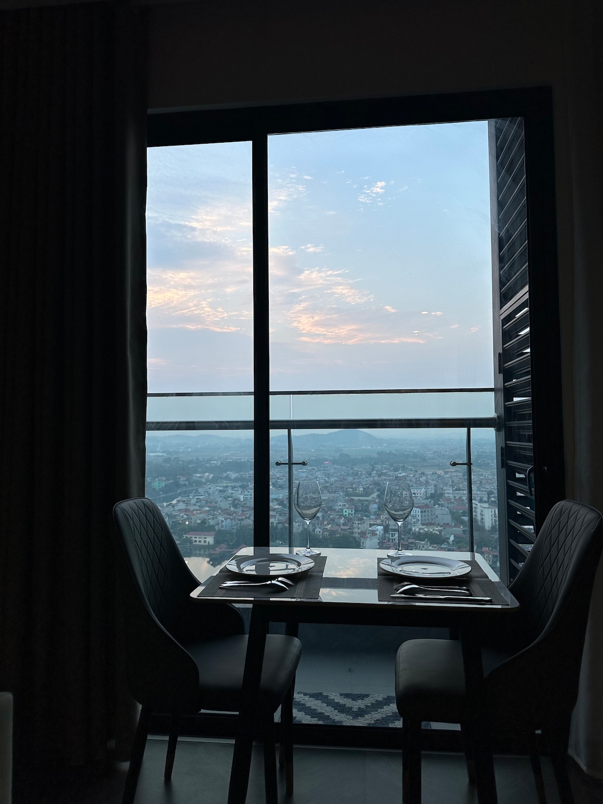 A dining area is positioned beside a large window offering expansive views of the city below. Two elegantly set plates and glasses rest on a small table, while the sky shows hints of sunset colors through the glass, framing a serene ambiance.