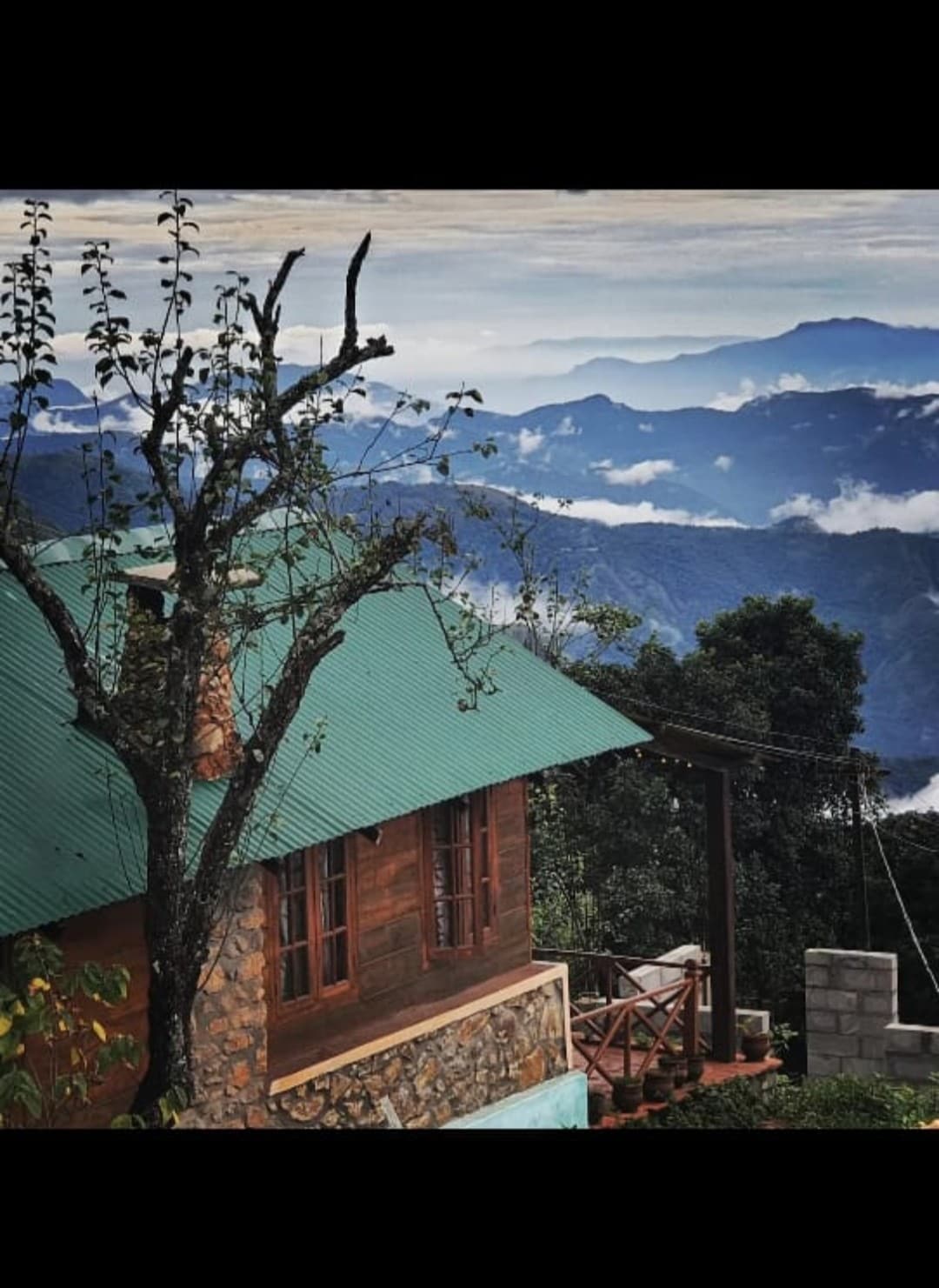 A rustic cabin with a green metal roof is nestled in the mountains, surrounded by trees and lush greenery. Rolling hills and misty clouds create a serene backdrop, enhancing the natural beauty of the location. A wooden deck with outdoor seating faces the scenic views.