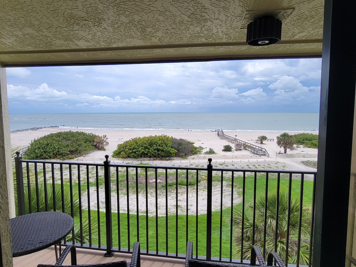 A private balcony overlooks a sandy beach and calm waters, with a wooden boardwalk leading into the distance. Lush greenery is visible along the shore, enhancing the serene coastal view. The black railing frames the scene, providing an inviting outdoor space.