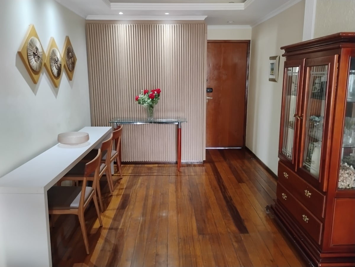 An entrance area features polished wooden flooring and soft lighting. A glass-top table is accompanied by two wooden chairs. A vase of red roses sits atop the table. A decorative cabinet is positioned against the wall, showcasing various items beneath its glass door.