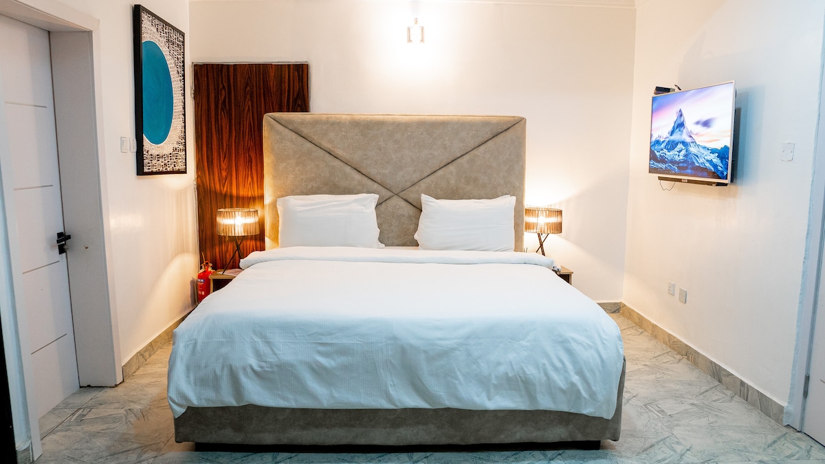 A spacious bedroom features a large bed flanked by two modern nightstands with lamps. A wall-mounted television is positioned across from the bed. Neutral tones are complemented by a piece of wall art, and textural variations are seen in the headboard and paneling.