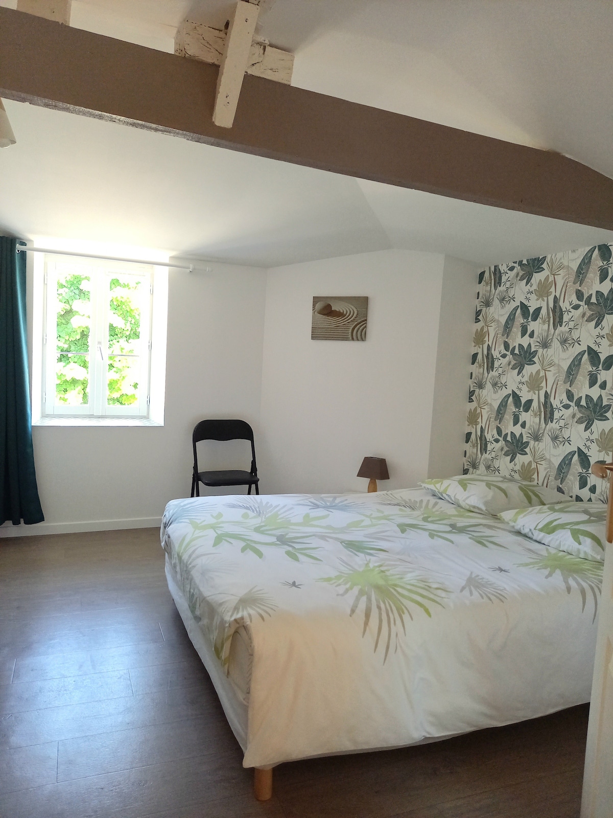 A bright sleeping area features a spacious bed adorned with a floral-patterned duvet. A folding chair is placed in the corner, next to a small lamp. Natural light filters through a window, complemented by green and white curtains. A decorative wall art piece adds a subtle touch.