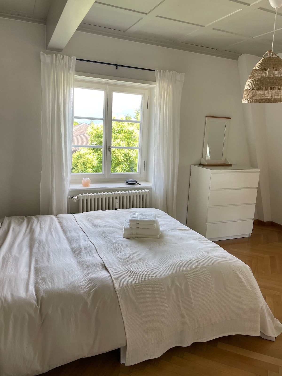 A serene bedroom features a spacious bed with neatly arranged towels on top. A window allows natural light to illuminate the room, complemented by soft white curtains. A light-colored dresser and a decorative mirror enhance the minimalist decor.