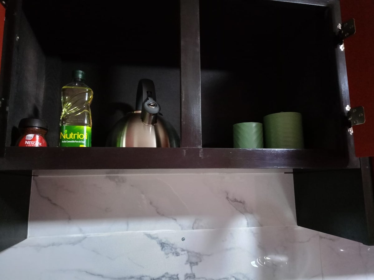 A kitchen cabinet is shown with two dark wood doors open. Inside, a silver kettle sits beside a bottle of cooking oil and two green cups. The countertop features a marble design, complementing the cabinetry.