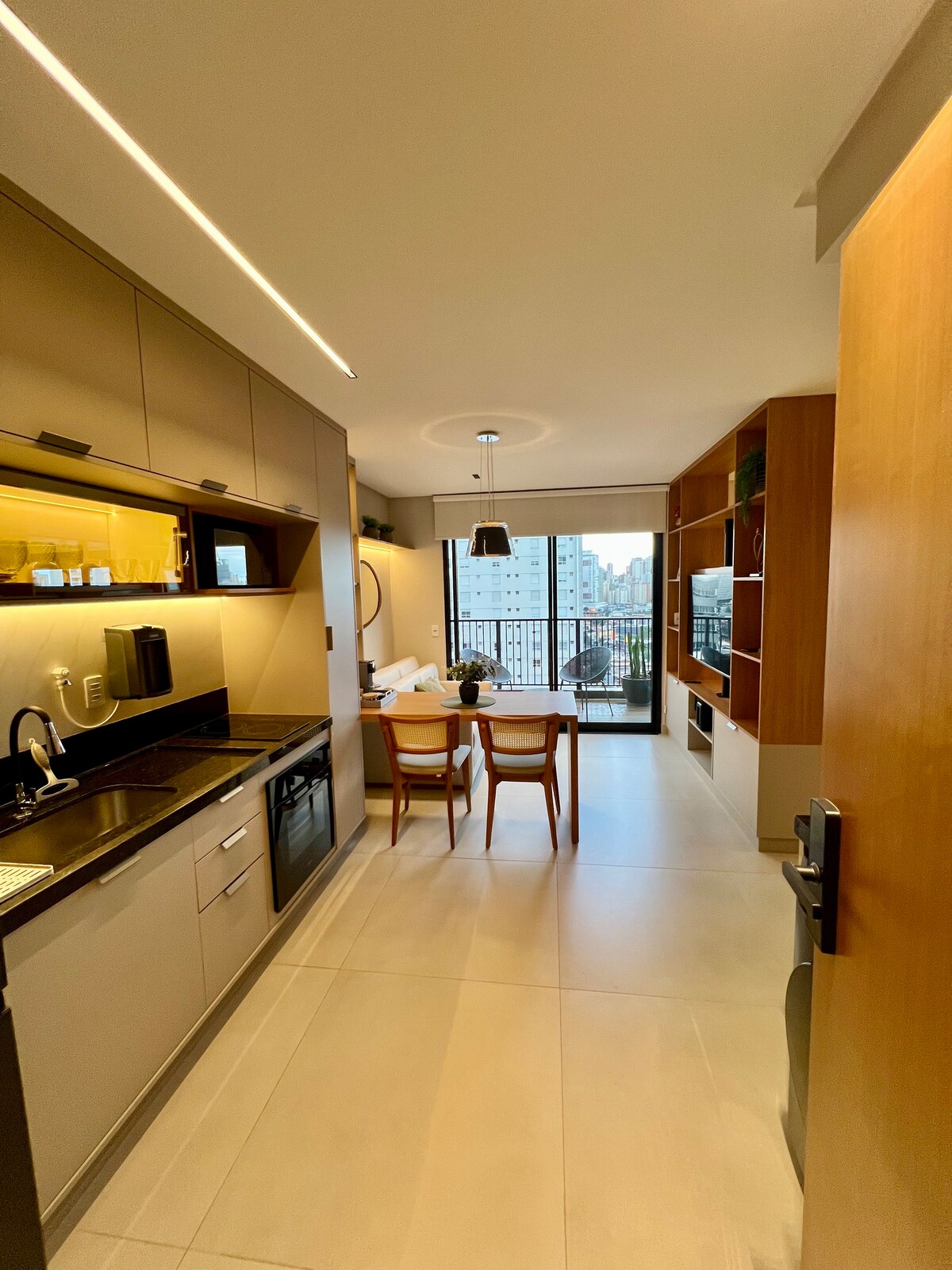 A modern kitchen area is depicted with sleek cabinetry and integrated appliances. A dining table for two is positioned near a large window, offering natural light and a view of the cityscape. The space is designed with clean lines and a neutral color palette.