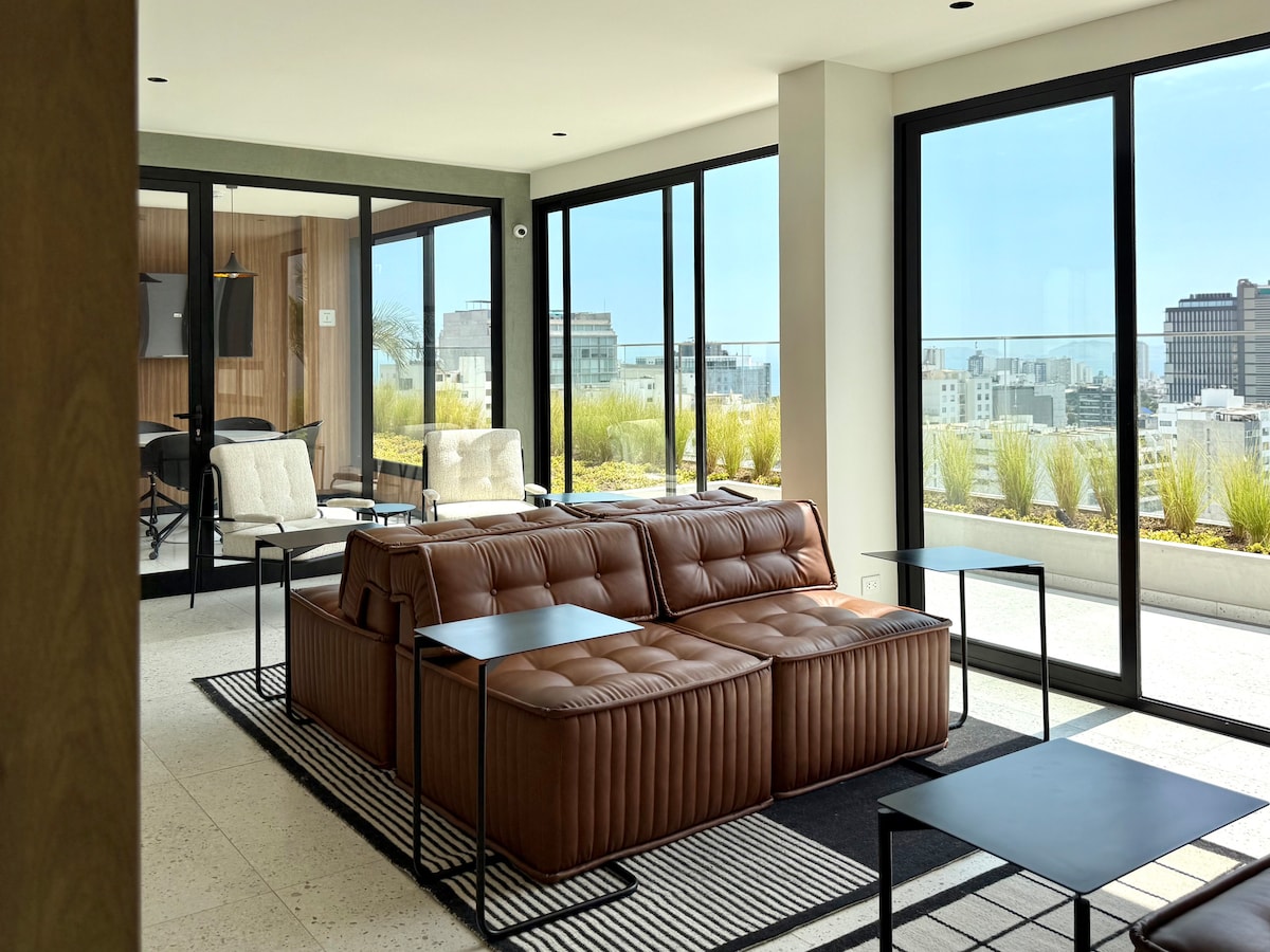A spacious living area is highlighted by a modern leather sofa, paired with sleek side tables. Floor-to-ceiling windows provide ample natural light, showcasing city views. A minimalist design is complemented by a neutral color palette and a patterned rug, creating a comfortable ambiance.