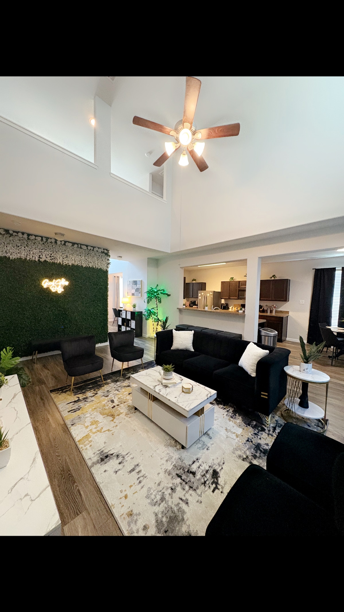 A spacious living area is adorned with modern furnishings, including a dark sectional sofa and accent chairs. A textured green wall adds a natural touch. A coffee table with decorative items is centered on a plush area rug, while a ceiling fan provides comfort.