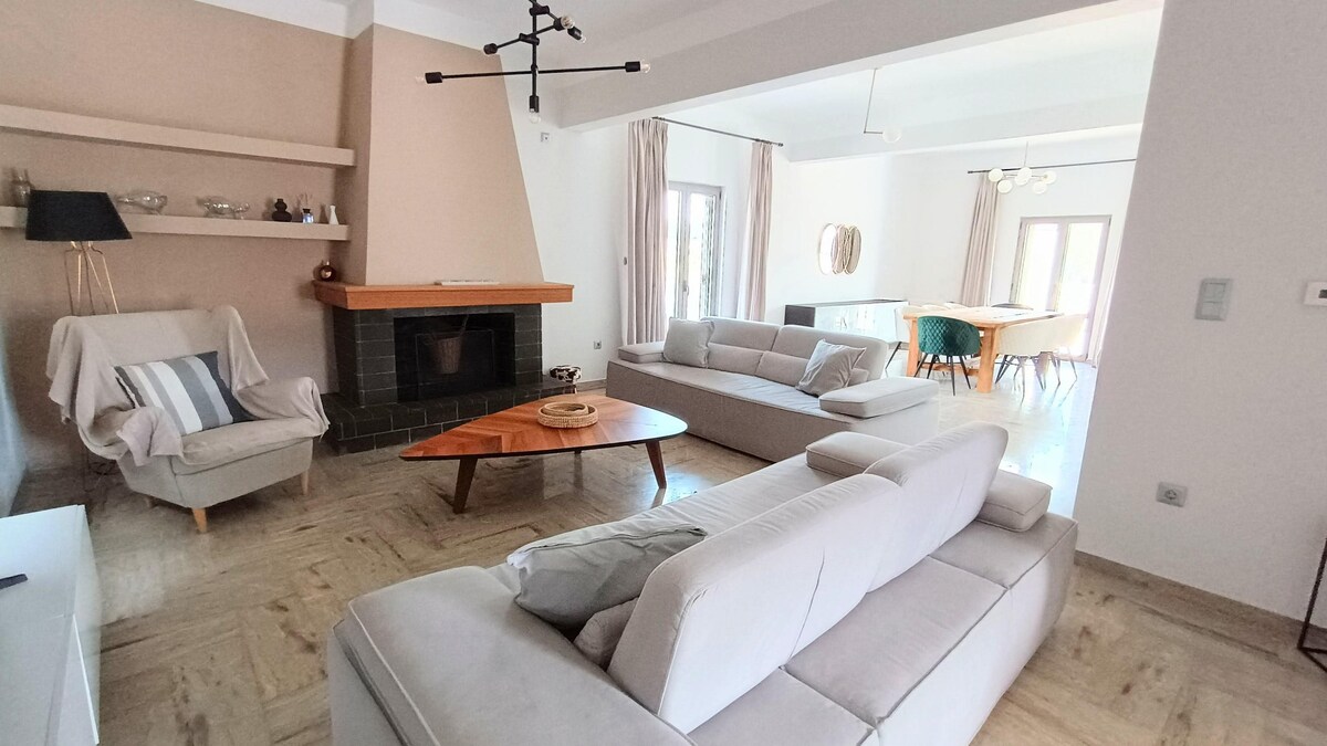 A welcoming living area features light-colored sofas arranged around a wooden coffee table. A fireplace is positioned against a soft beige wall, contributing to a cozy ambiance. Natural light filters through large windows, illuminating the space, which connects to a dining area in the background.