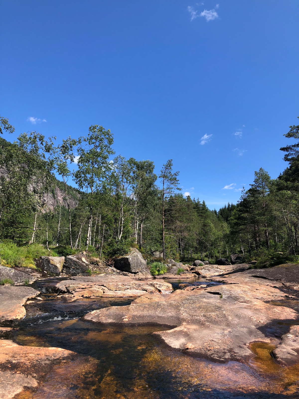 A clear blue sky is overhead, accompanied by scattered clouds. A shallow river flows over smooth, large stones, surrounded by lush greenery and tall trees. The rocky riverbed reflects the sunlight, creating a serene and inviting natural setting.