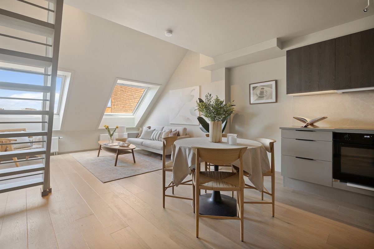 A bright living area is showcased with natural light streaming through triangular windows. A comfortable sofa is paired with a round dining table set for four. The kitchen area features modern cabinetry, while a decorative plant adds a touch of greenery to the inviting space.