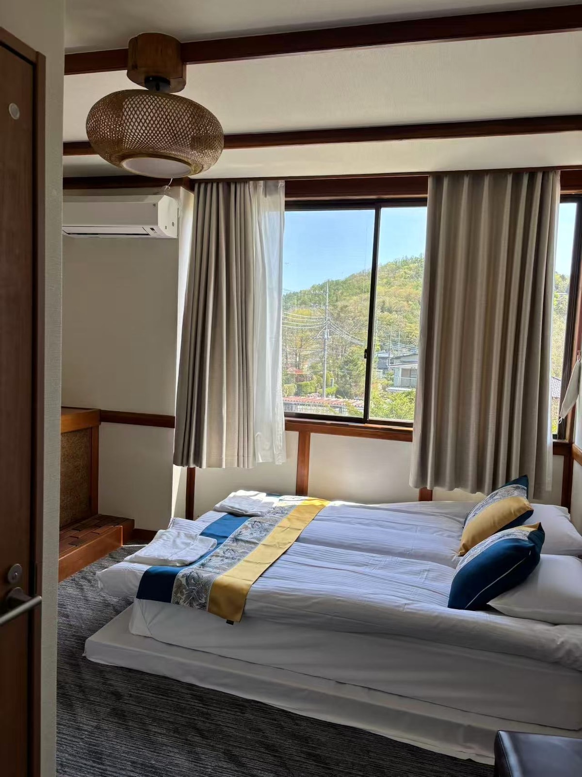 A cozy sleeping area is featured with a spacious bed dressed in white linens and decorative pillows. Large windows allow natural light to fill the room, complemented by sheer curtains. A woven light fixture hangs from the ceiling, enhancing the overall ambiance.