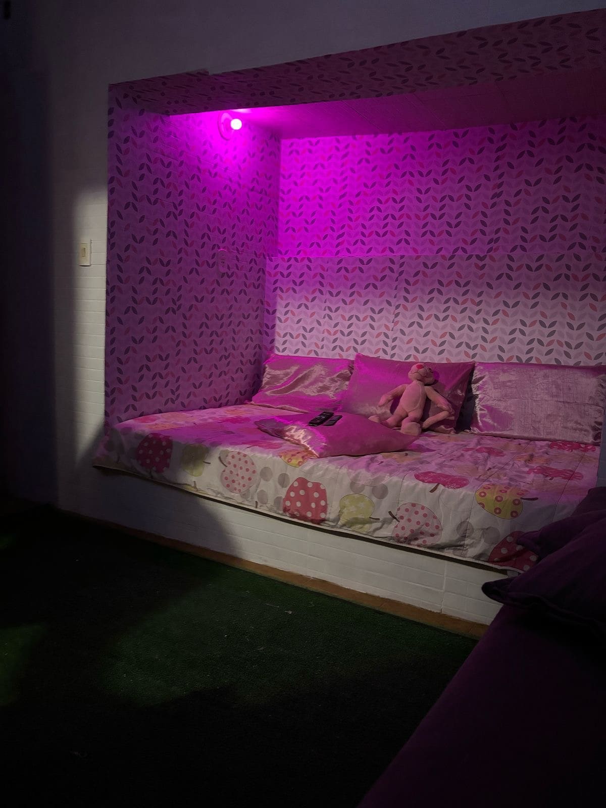A cozy sleeping nook is displayed with soft bedding in floral patterns. A plush toy sits among vibrant pink pillows. The space is softly illuminated by purple lighting, creating a calming atmosphere. The wall is decorated with a whimsical patterned wallpaper.