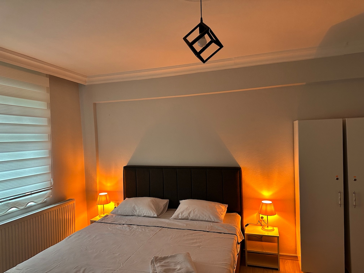 A well-appointed bedroom features a spacious bed dressed in crisp white linens. Warm light emanates from two bedside lamps, creating a soft glow. A tall wardrobe is positioned against a neutral wall, while large windows with sheer coverings invite natural light into the space.