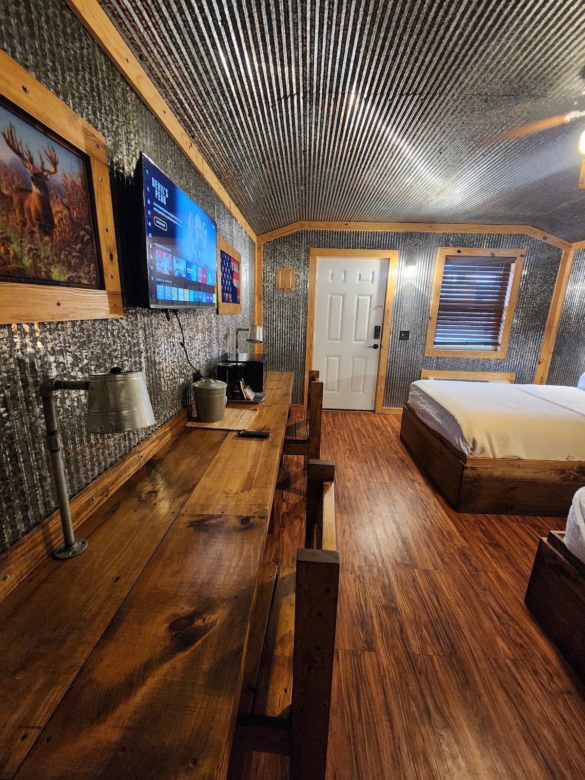 A rustic room is characterized by metal walls and wooden accents. Two queen beds are positioned beside a desk made of wood, which features a lamp and a small coffee station. A wall-mounted TV is visible, along with a door leading outside.