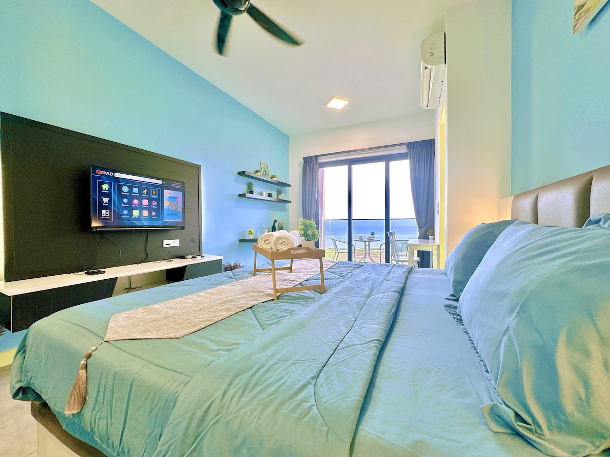A bright bedroom features blue walls and a spacious bed adorned with soft linen. A flat-screen TV is mounted on the wall, and a small table holds neatly rolled towels. Large windows allow natural light to fill the room, offering a view of the outdoors.