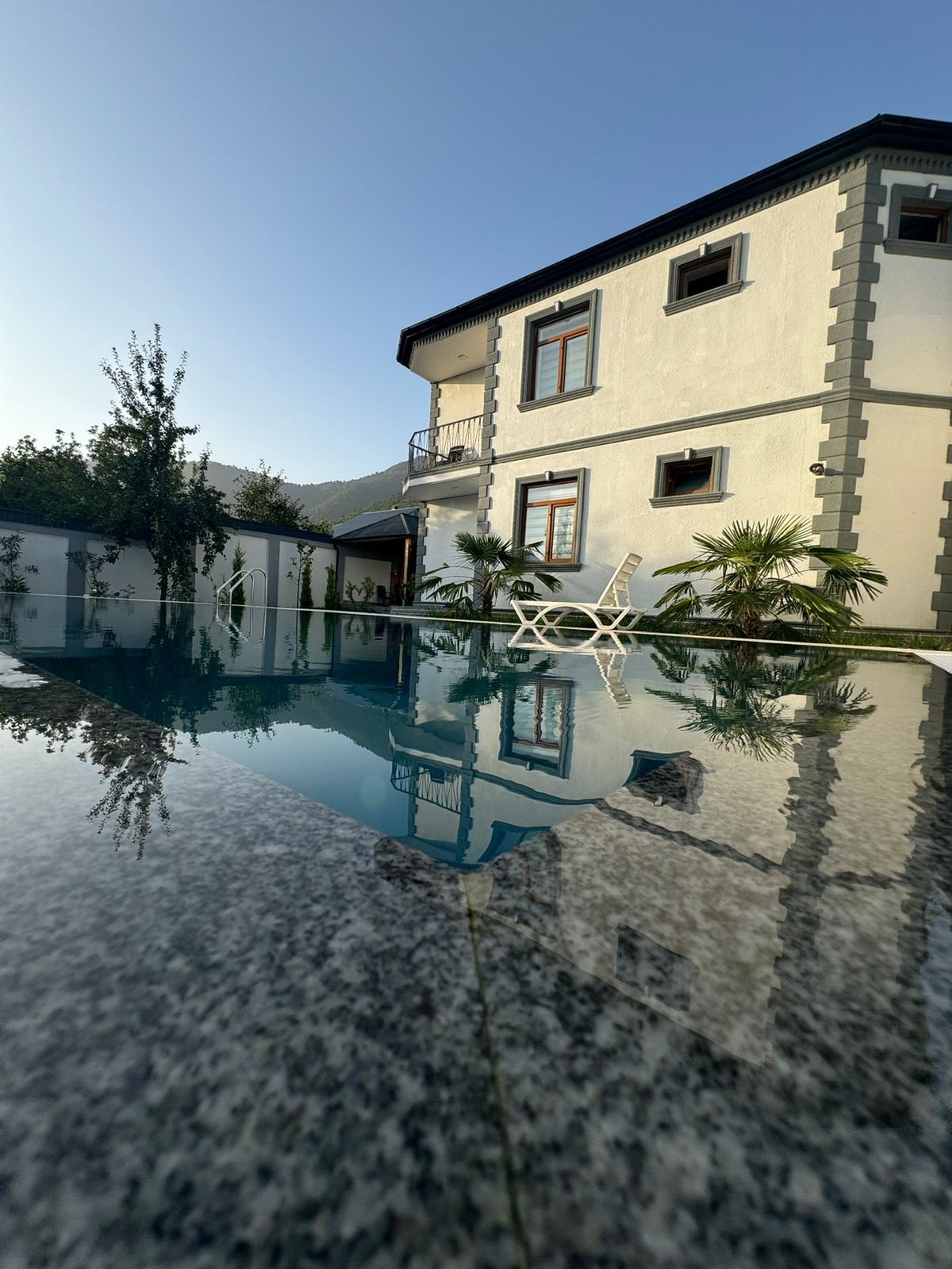 A serene outdoor setting features a shimmering pool reflecting the clear sky. The well-maintained exterior of a two-story house is visible, flanked by lush greenery and palm plants. Comfortable lounge chairs are positioned around the pool area, inviting relaxation.