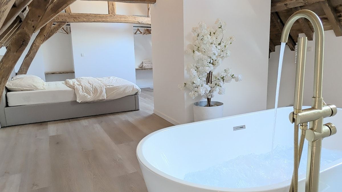 An airy room features exposed wooden beams, creating a charming architectural detail. A white freestanding bathtub includes water gently flowing from its faucet. A spacious bed is positioned in the background, complemented by a minimalist decor and a floral arrangement in a white pot nearby.