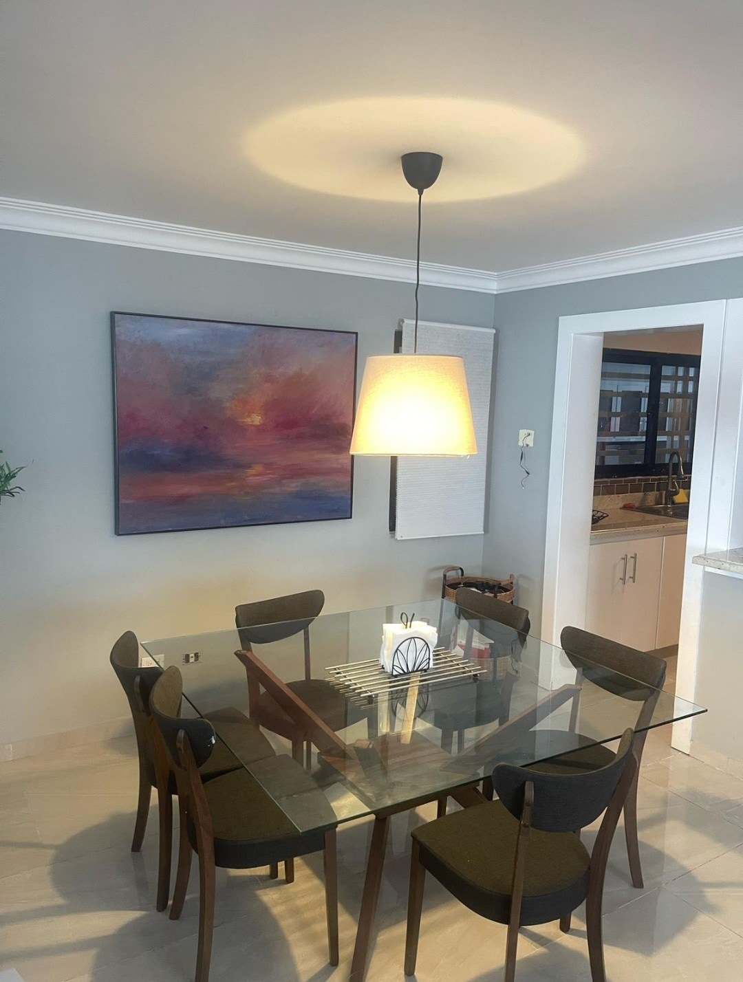A modern dining area features a glass table surrounded by six dark wooden chairs. A warm pendant light hangs above, illuminating a vibrant piece of artwork on the wall, which showcases abstract colors. Natural light can be seen coming from the adjacent kitchen area.