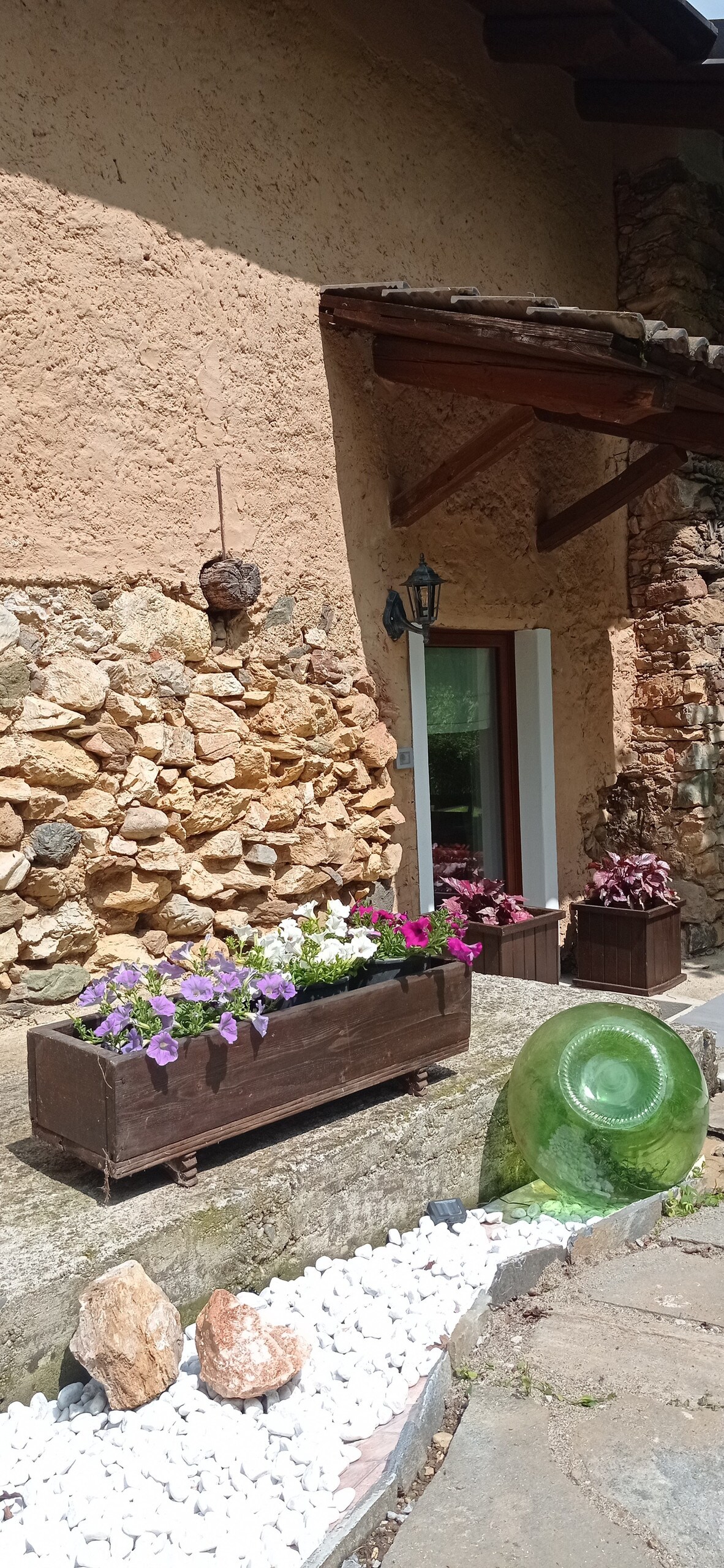 The entrance area features rustic stone walls and wooden accents. A wooden planter filled with colorful flowers is placed beside the door, while a large green decorative glass piece and white pebbles create an inviting pathway.