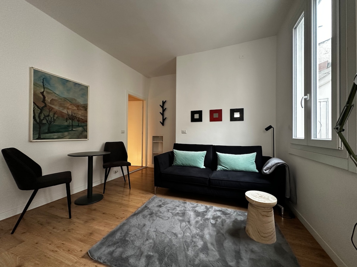 A bright living area is presented, featuring a comfortable black sofa paired with light green cushions. A small round table is set with two chairs, and a light rug covers the wooden floor. Art pieces adorn the walls, while natural light filters through the windows.