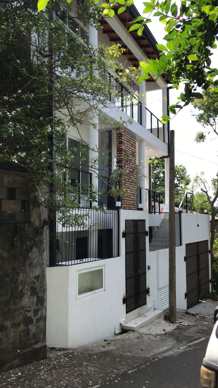 The exterior of the apartment building is displayed, featuring a multi-level structure with a blend of brick and white walls. Balconies provide outdoor space, while tree branches and foliage frame the scene. A pathway leads to the entrance, bordered by a stone wall.