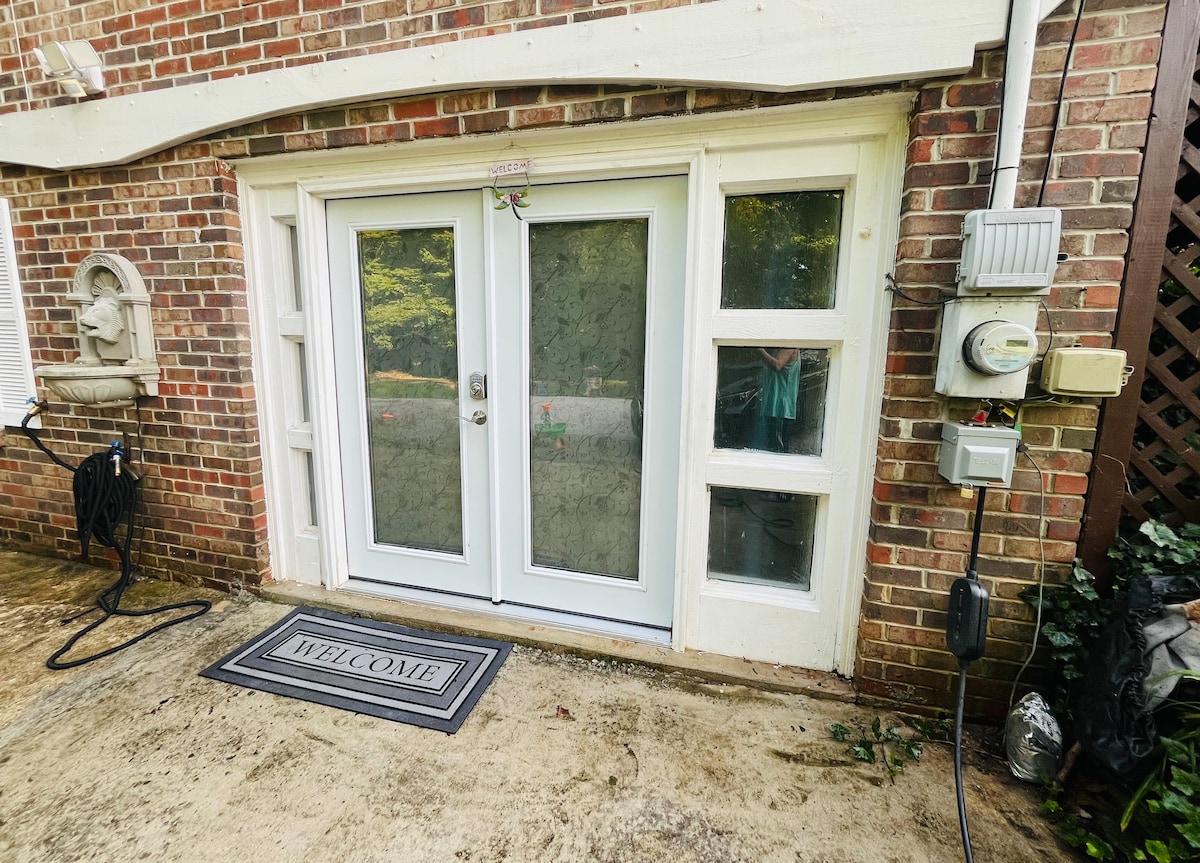 The entrance features a set of modern double glass doors framed by brick, allowing natural light to brighten the space. A welcome mat lies in front, and utility meters are visible on the side. Lush greenery surrounds the entrance, enhancing the inviting appearance.