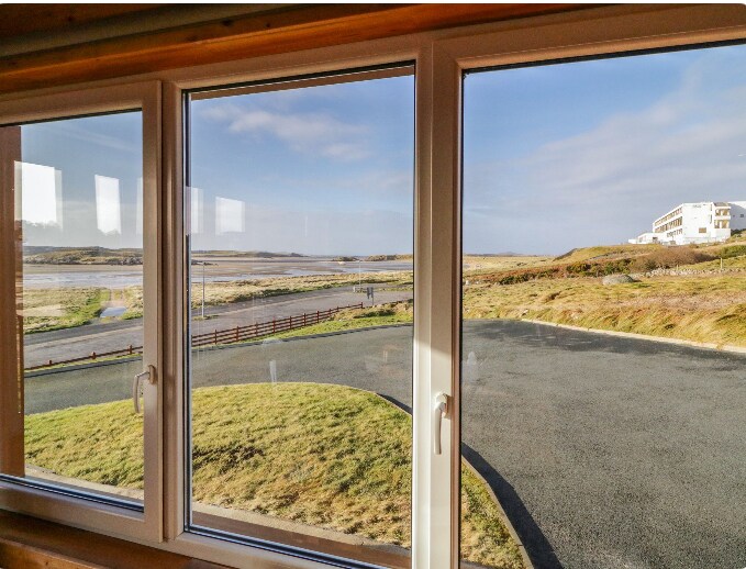 A spacious window offers a view of the tranquil coastal landscape, showcasing lush green grass and a winding road leading to Bunbeg Beach. The sky is clear with soft, natural light illuminating the scene.