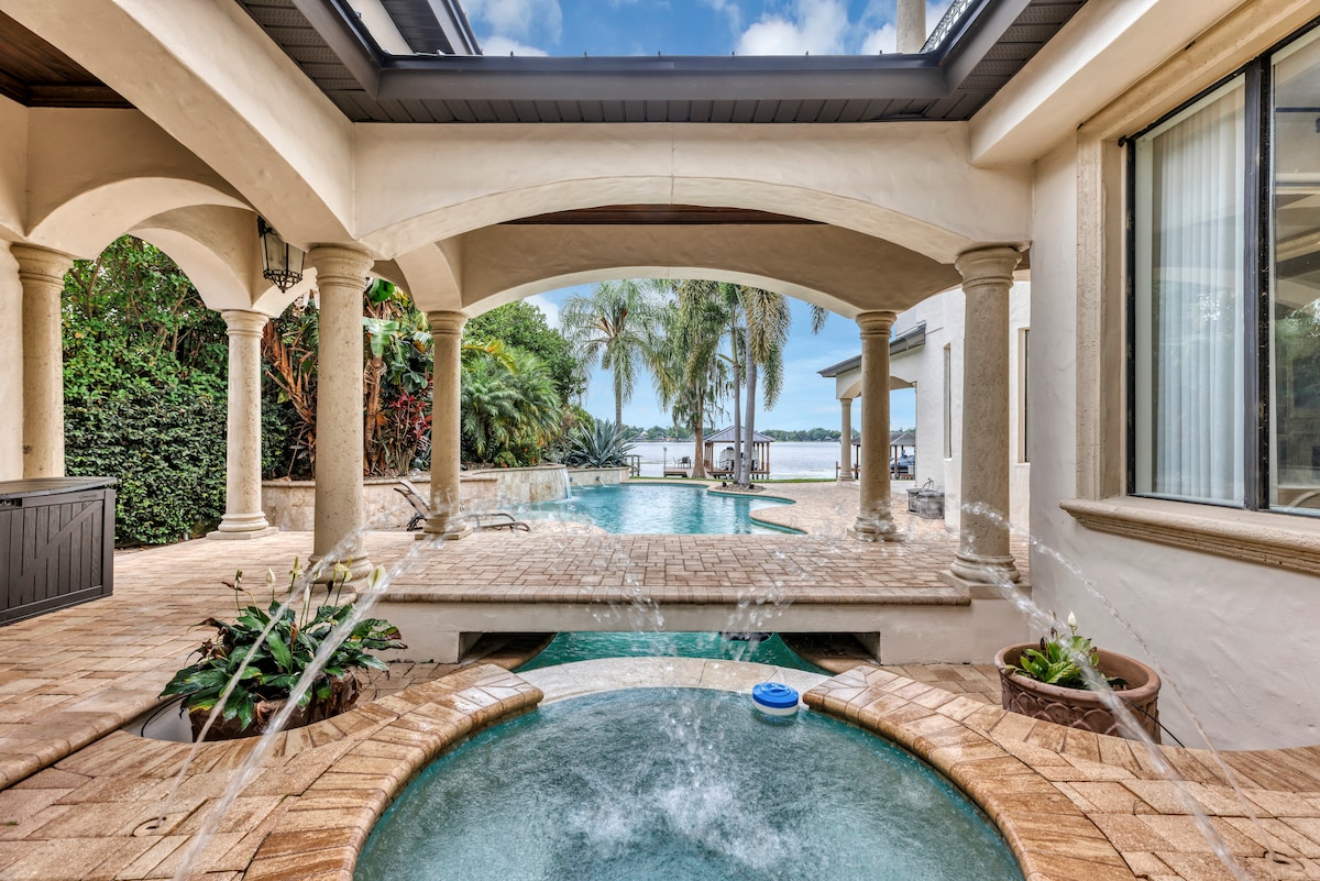 An outdoor patio area features elegant arches and stone columns, overlooking a sparkling pool. Water fountains create a serene ambiance, while palm trees surround the space. The backdrop reveals a peaceful lake, contributing to the tranquil environment.