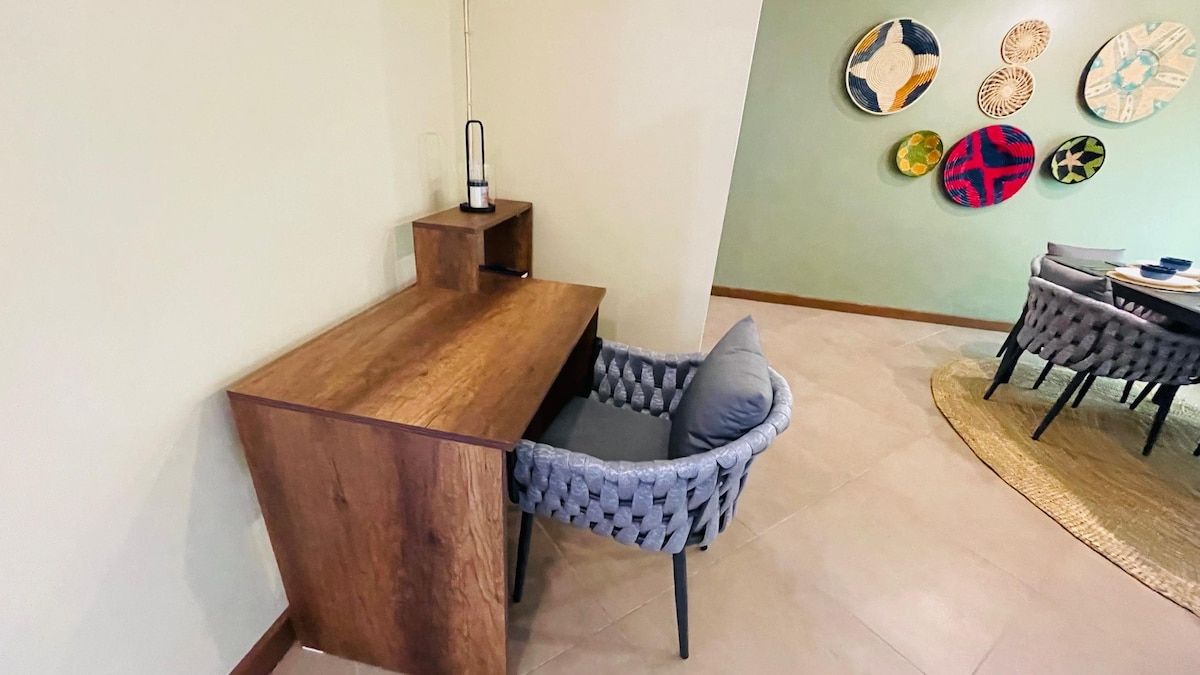 A well-designed work station features a wooden desk accompanied by a comfortable chair. The wall displays decorative art pieces, adding a touch of color to the neutral surroundings. The floor is tiled, enhancing the modern appeal of the space.