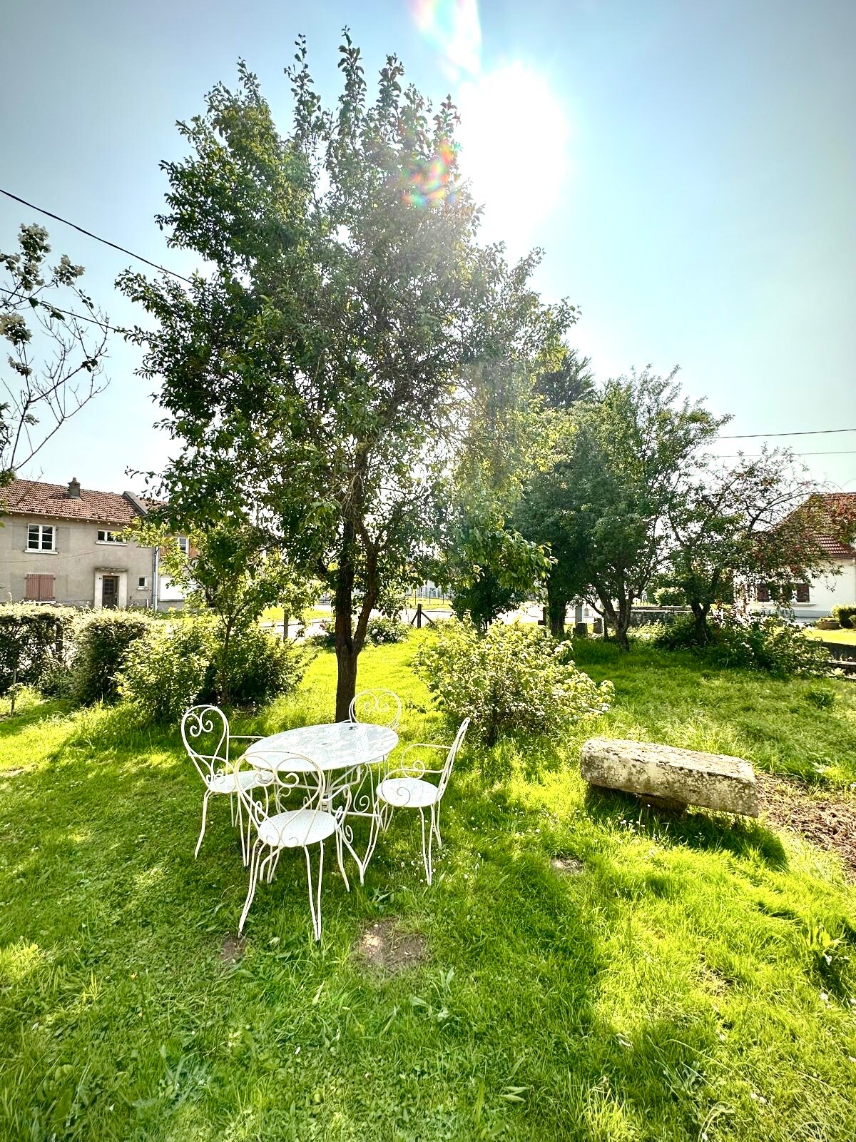 A small outdoor seating area is framed by greenery, featuring a round table with intricate metalwork and two matching chairs. Sunlight casts a warm glow over the lush grass, accentuating the surrounding trees and inviting atmosphere. A stone can be seen partly hidden in the landscape.