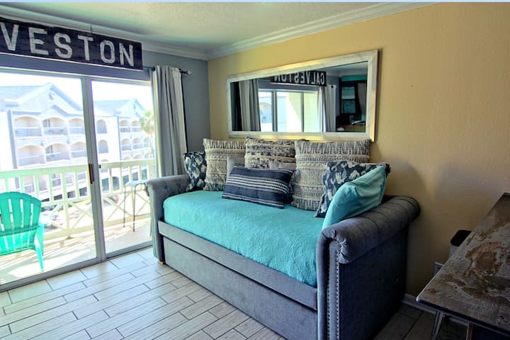 Bright coastal living space with a comfy daybed, beachy accents, and sliding doors opening to a private balcony. Perfect spot to relax, read, or enjoy the ocean breeze just steps from the beach.