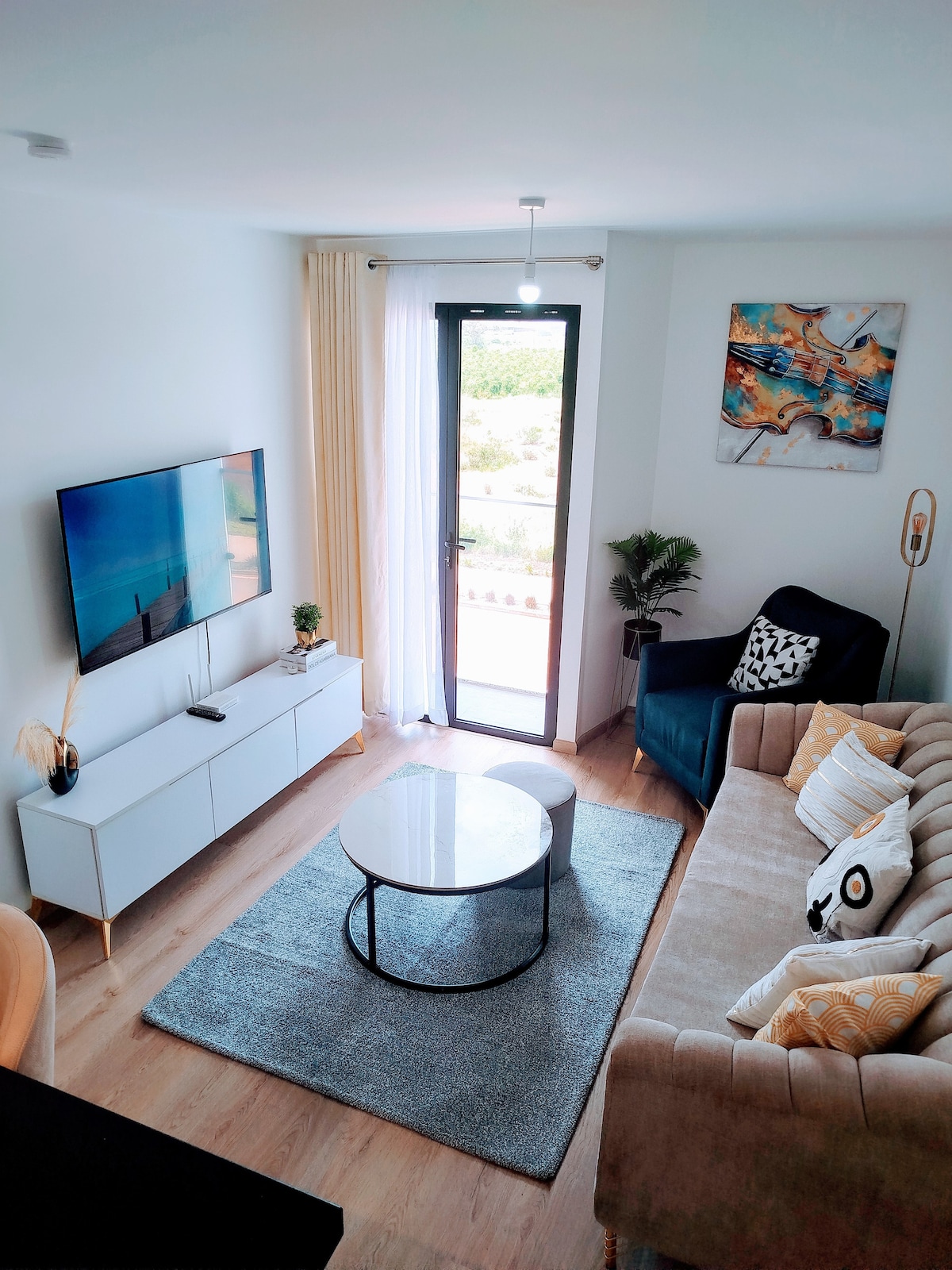 A modern living area features a light-colored sofa and a dark armchair, complemented by a round coffee table. A flat-screen television is mounted on the wall, while natural light enters through a glass door that opens to a balcony. Soft textures enhance the space.