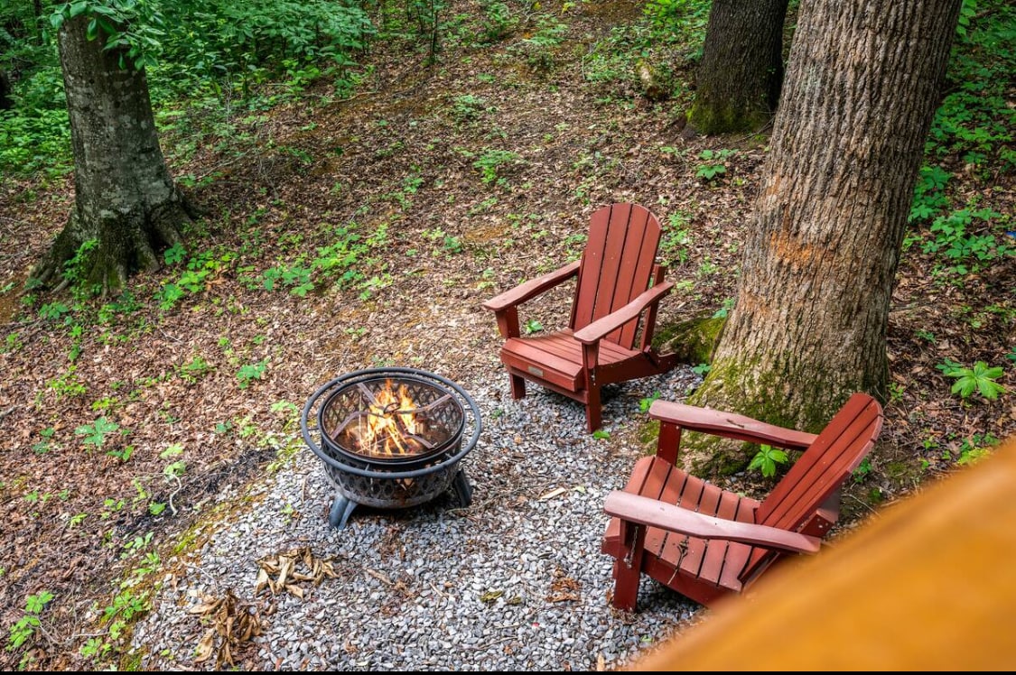 A cozy outdoor fire pit area features two wooden Adirondack chairs set on a gravel base. The fire pit, surrounded by stones, holds a warm, glowing fire, while lush green foliage from nearby trees provides a serene backdrop.