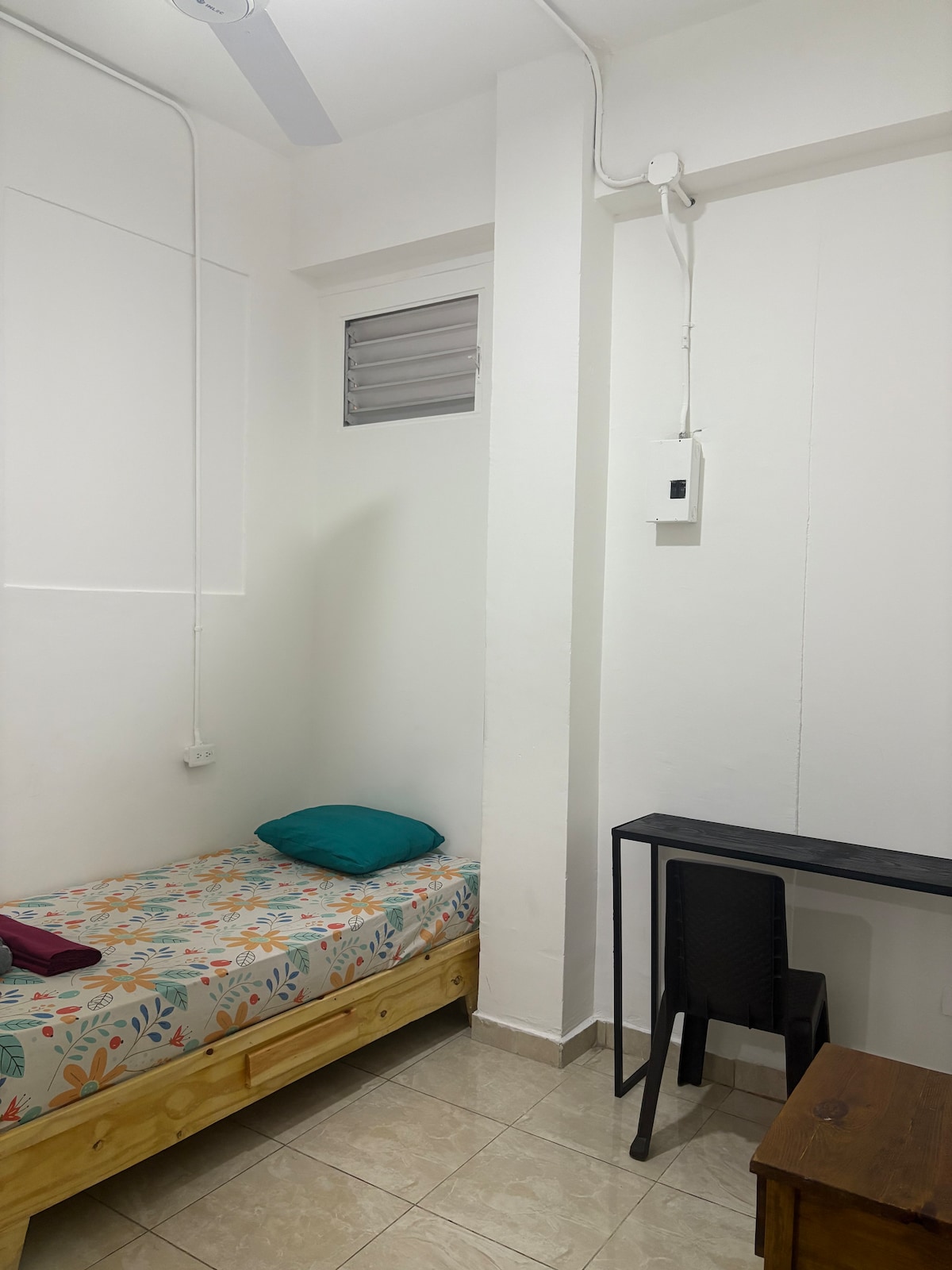 A simple room setup is presented, featuring a single bed with a floral-patterned blanket and a teal cushion. A small wooden desk and chair are positioned nearby, providing workspace. Natural light enters through a small window, enhancing the bright and clean white walls.