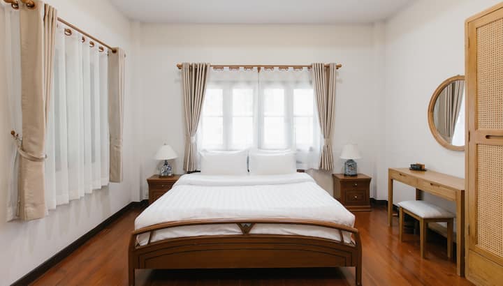 Dhomë gjumi autentike në Chiang Mai me mobilje prej druri teak në stil Lanna dhe thekse prej ratani. Ka një tavolinë makijazhi minimaliste, pasqyrë makijazhi rrethore dhe dritë natyrale të butë me shkëlqim të bollshëm. Një parajsë paqësore dhe estetike për blogerët e udhëtimeve dhe ata që kërkojnë mirëqenie.