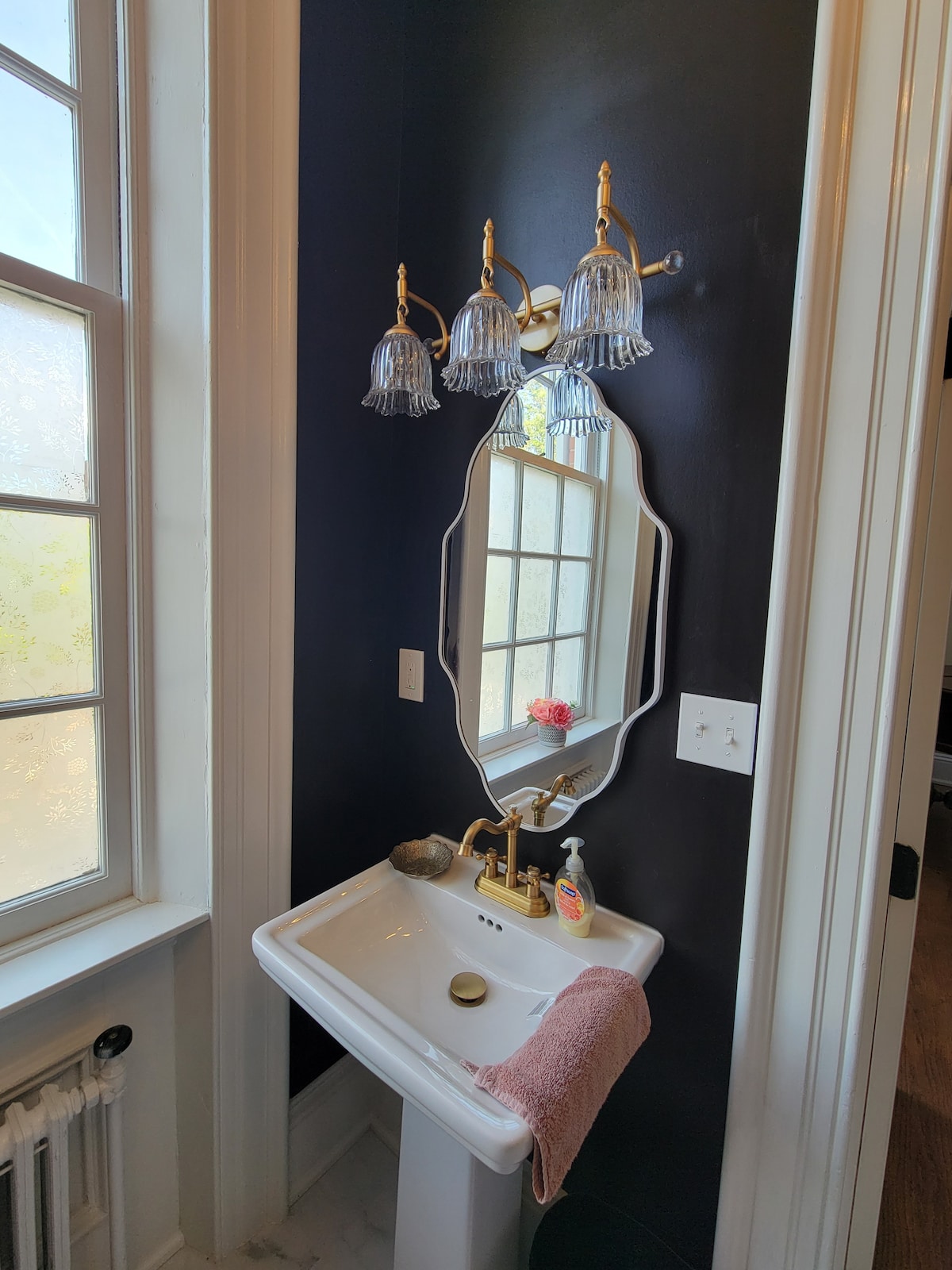A classic bathroom features a white pedestal sink with a gold faucet and a shaped mirror above it. The walls are painted dark blue, accentuated by a decorative four-light fixture. A pink towel is draped over the sink edge, and natural light filters through the windows.