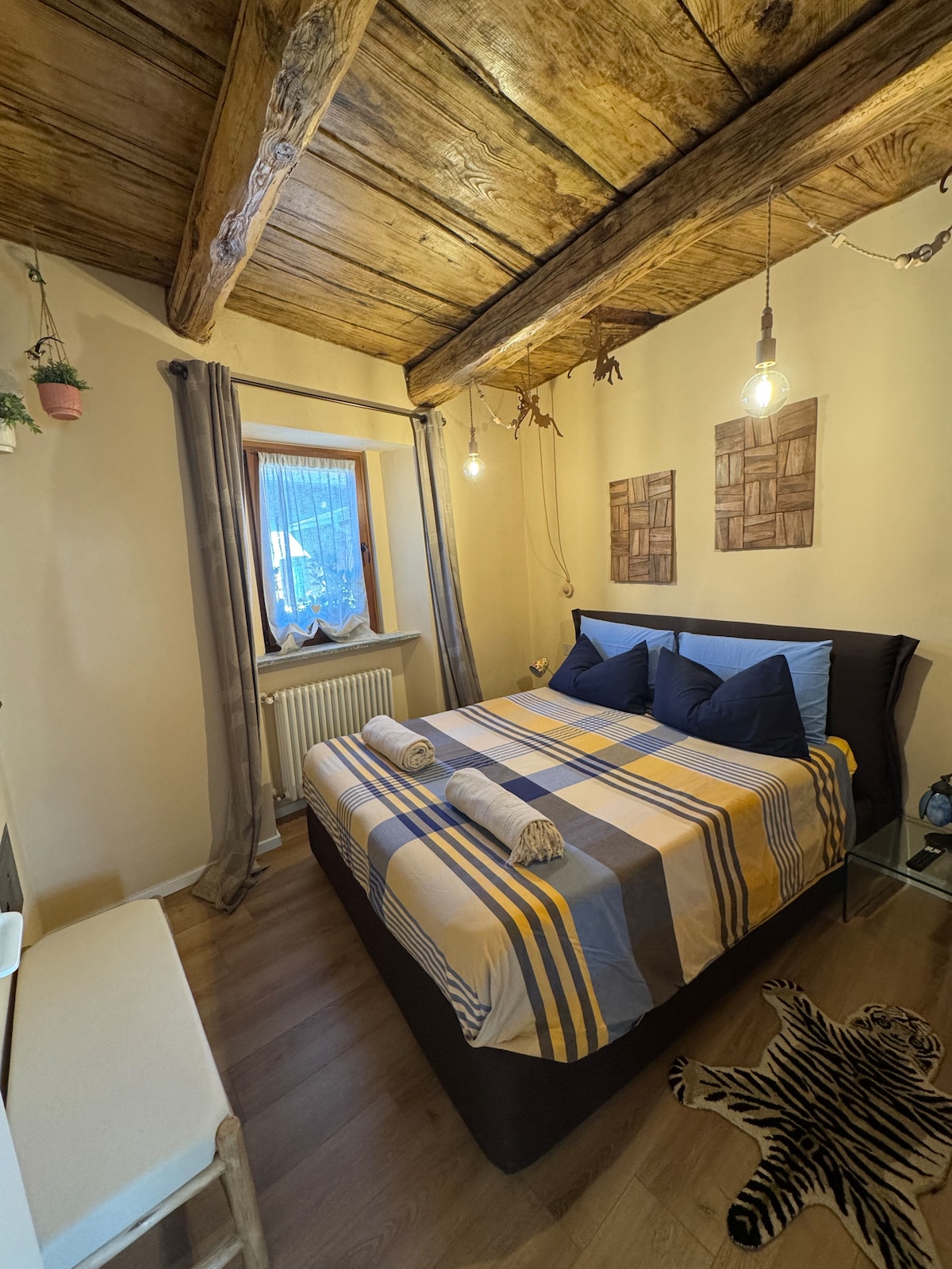 A cozy bedroom featuring a double bed with a checkered blanket and two rolled towels on top. Natural light filters in through a window, complemented by soft curtains. Exposed wooden beams line the ceiling, and decorative elements can be seen on the walls.