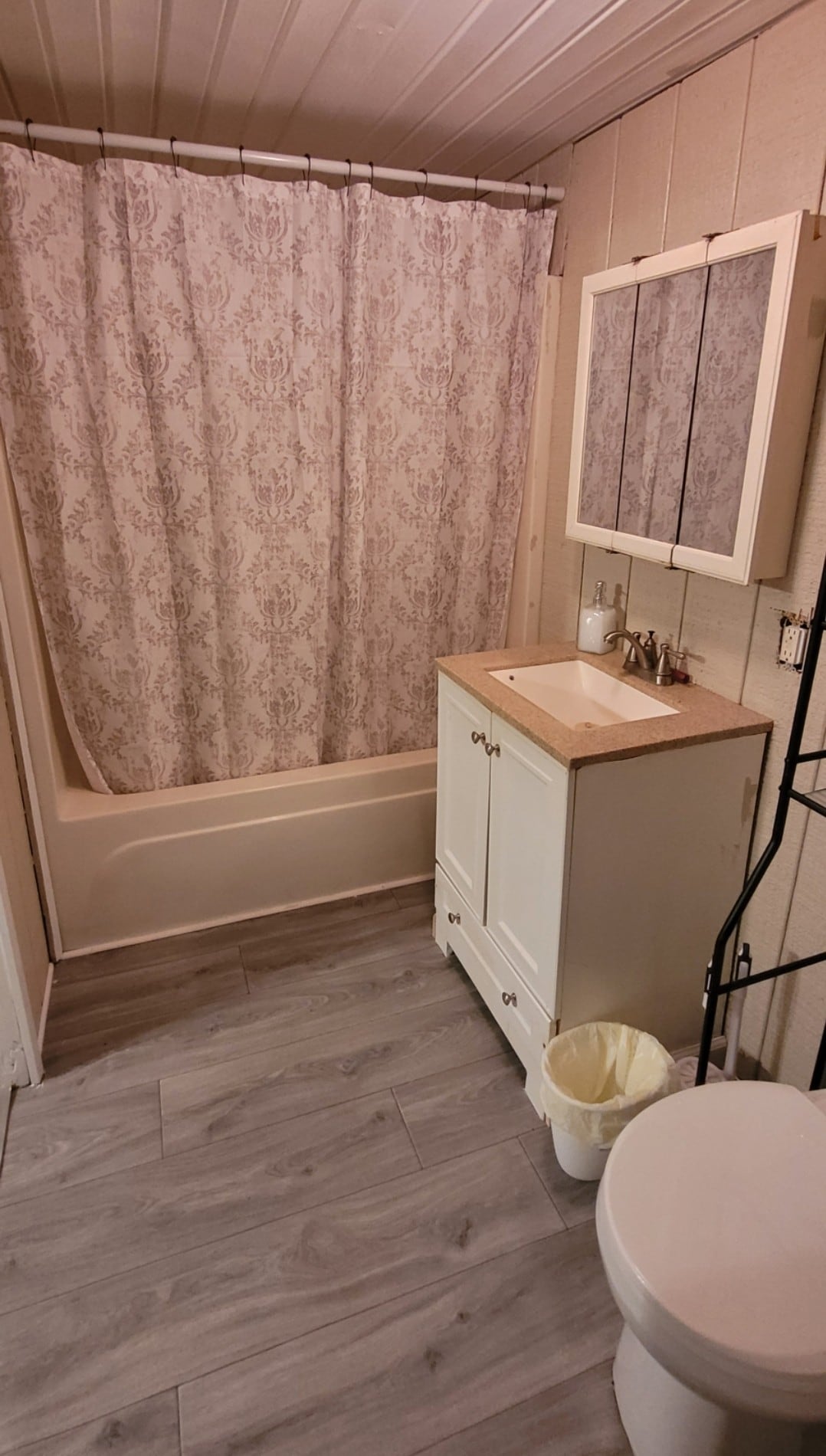 A compact bathroom features a clean design with a bathtub and shower combination. Light-colored walls are complemented by a textured shower curtain. A vanity with a light wooden countertop and sink is positioned beside a mirror, providing functional space alongside a white toilet.