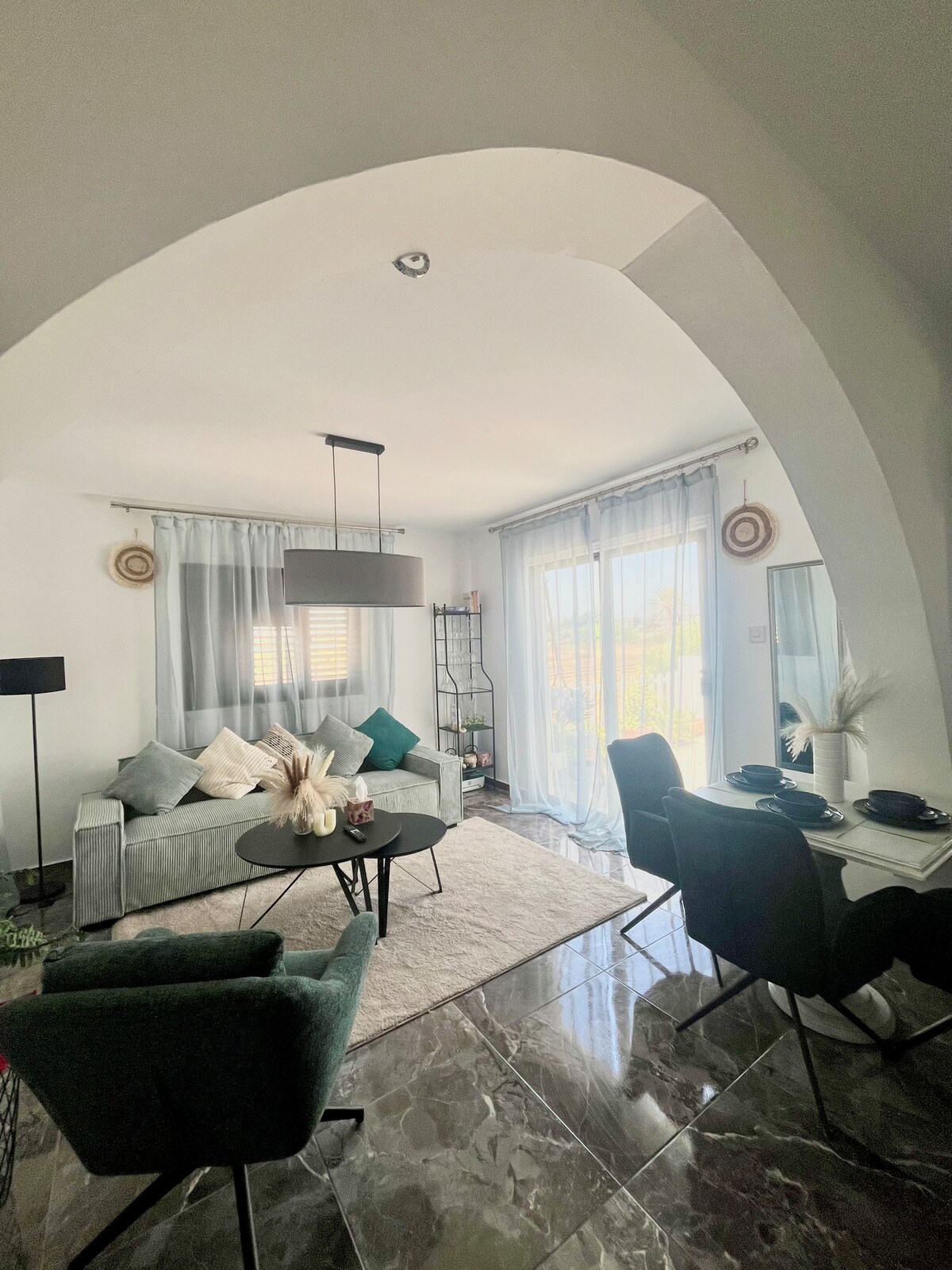 An inviting living area is characterized by large windows that allow natural light to fill the space. A comfortable grey sofa is complemented by soft pillows. A circular black table and two green chairs provide a cozy spot for dining, with a decorative shelving unit in the background.