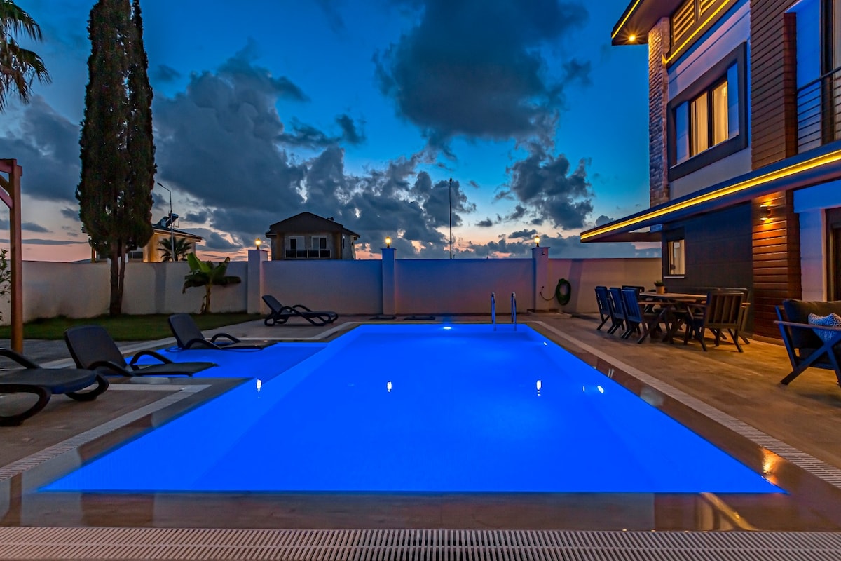 A half-lit pool area showcases a shimmering blue water surface under a twilight sky. Surrounding the pool, several lounge chairs are positioned for relaxation, while a nearby dining table indicates the potential for outdoor meals.