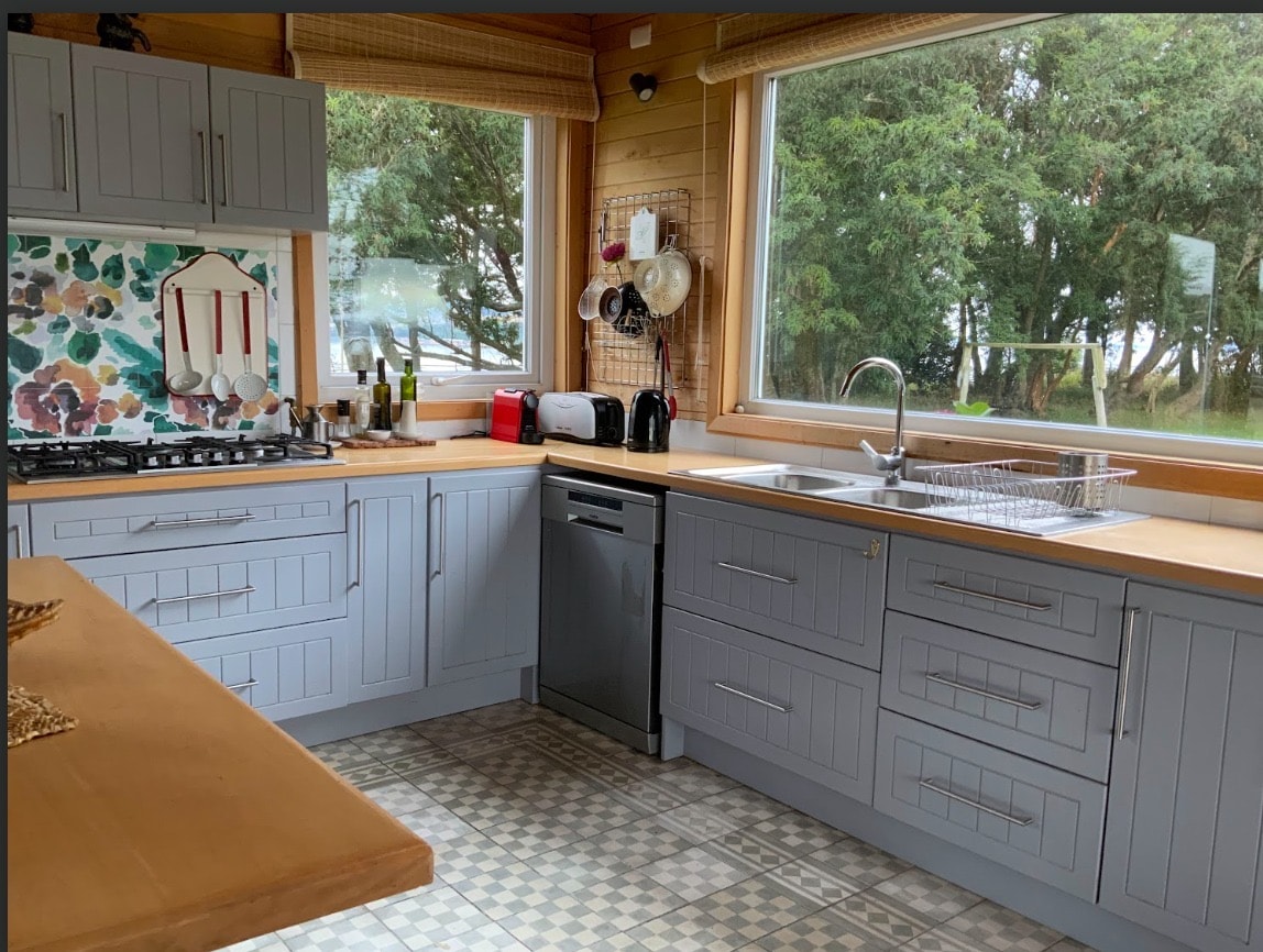 A well-equipped kitchen features gray cabinets and ample counter space. Large windows provide views of the surrounding greenery, while a sink and dishwasher are positioned near the cooking area. Colorful wall tiles add a touch of vibrancy to the space.