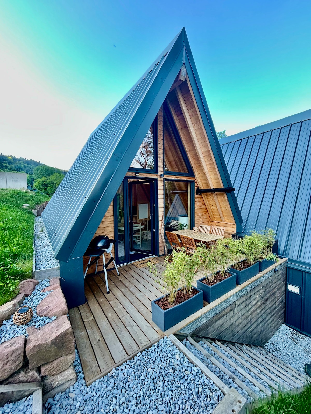 A modern A-frame structure showcases a wooden terrace with a dining set, positioned among planters filled with greenery. Glass doors allow light to enter, leading into the interior. The exterior features a combination of wood and metal, set against a backdrop of lush grass.