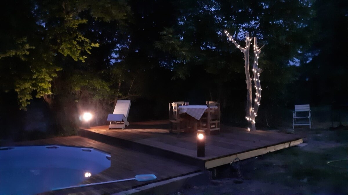 A wooden deck is illuminated softly at twilight, featuring a small swimming pool and stylish seating. A table set for dining is surrounded by cozy chairs, while nearby trees are adorned with warm string lights, enhancing the serene outdoor space.