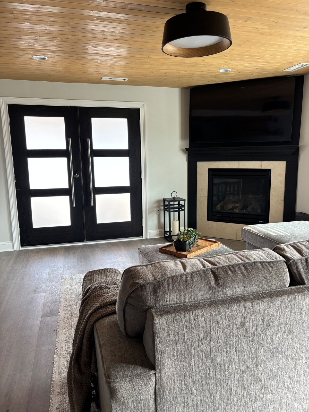 A contemporary living area is highlighted by large double doors with a frosted glass design, complementing a mounted television above a sleek gas fireplace. A comfortable sectional sofa is adorned with a textured throw blanket, while a small coffee table holds a decorative plant and candle.