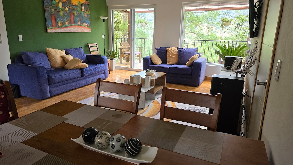 A spacious living area features two blue sofas and a coffee table set on a patterned rug. Large windows provide natural light and views of the surrounding greenery. A wooden dining table is set with decorative items, enhancing the inviting atmosphere.