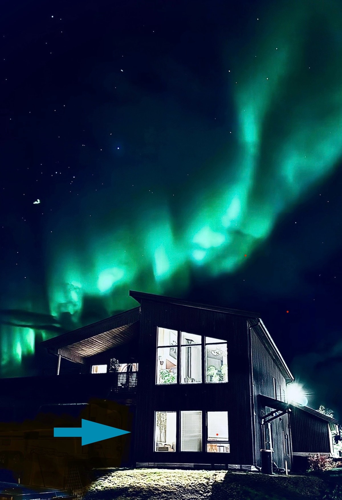 A modern cabin is illuminated under a vibrant display of the northern lights, with bright green hues dancing across the night sky. Large glass windows offer a glimpse of the indoor space, emphasizing a connection with the stunning natural surroundings.