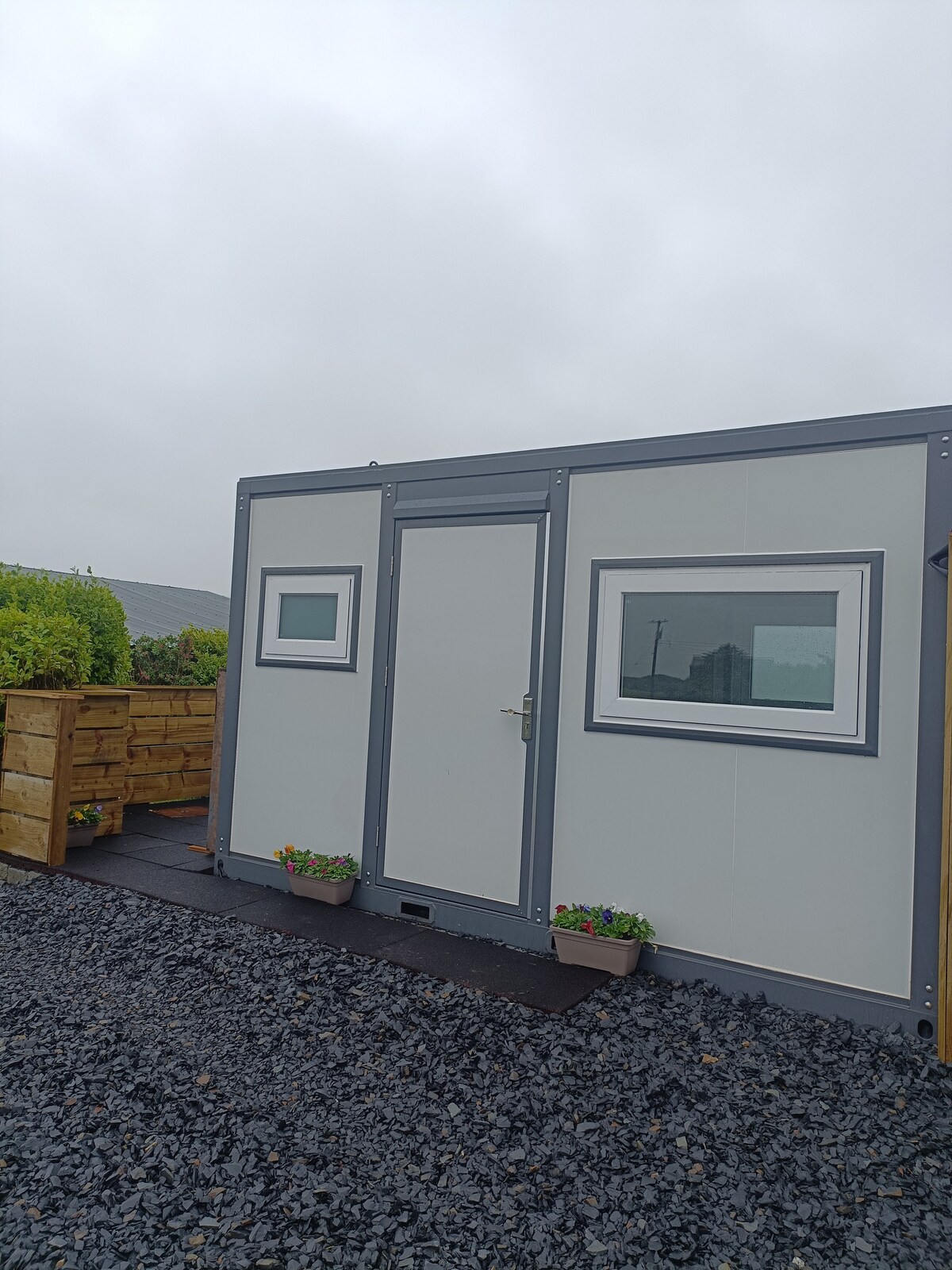 A modern cabin exterior is shown, featuring a main entrance door and two small windows outlined in contrasting trim. Planters with vibrant flowers are positioned on a wooden deck, leading to a gravel pathway. The surrounding area features greenery under a cloudy sky.