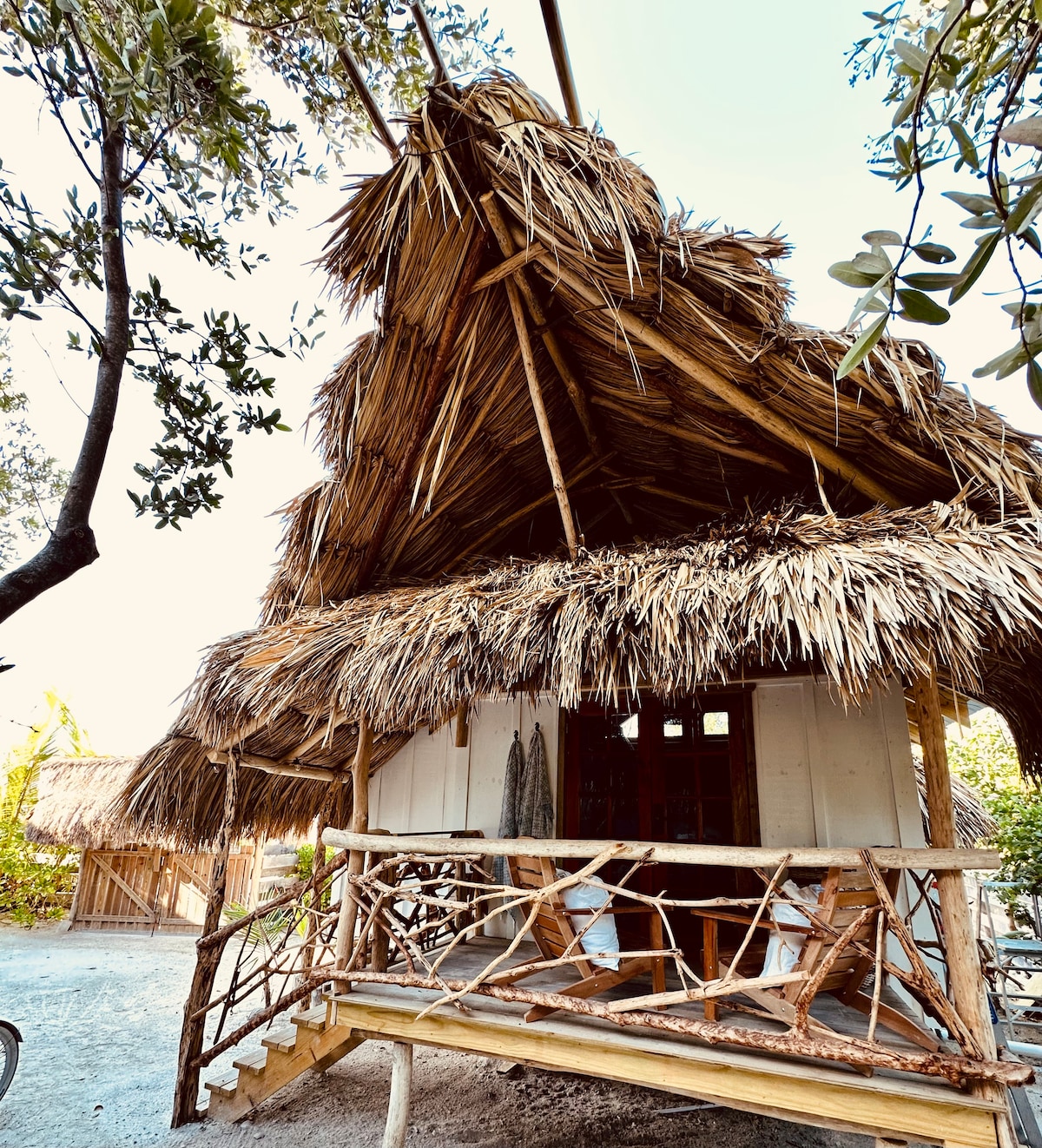 A charming hut with a thatched roof stands among greenery, showcasing natural materials. The structure features a spacious porch supported by wooden railings, with rustic decor elements visible. Surrounding trees offer shade, enhancing the peaceful environment of the property.