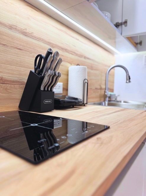 A modern kitchen countertop features a sleek black induction cooktop beside a stylish knife block filled with various utensils. A roll of paper towels is positioned nearby, with soft lighting illuminating the warm wood grain of the surface.